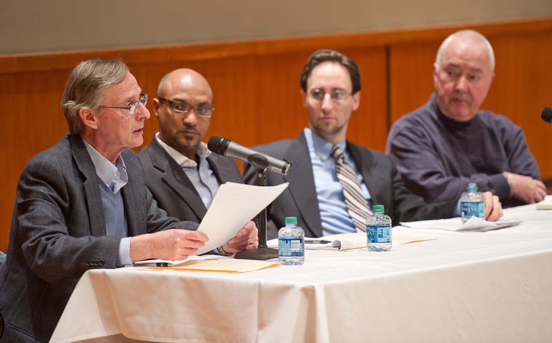 A panel of four men sit at a white tablecloth table with a microphone, papers and bottles of water.