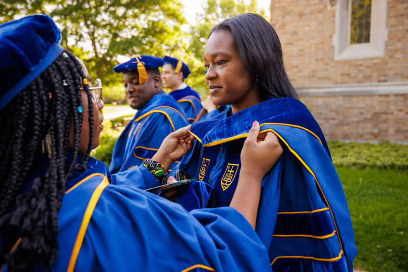 Woman receiving assistance with ceremonial wardrobe prior to commencement ceremonies.