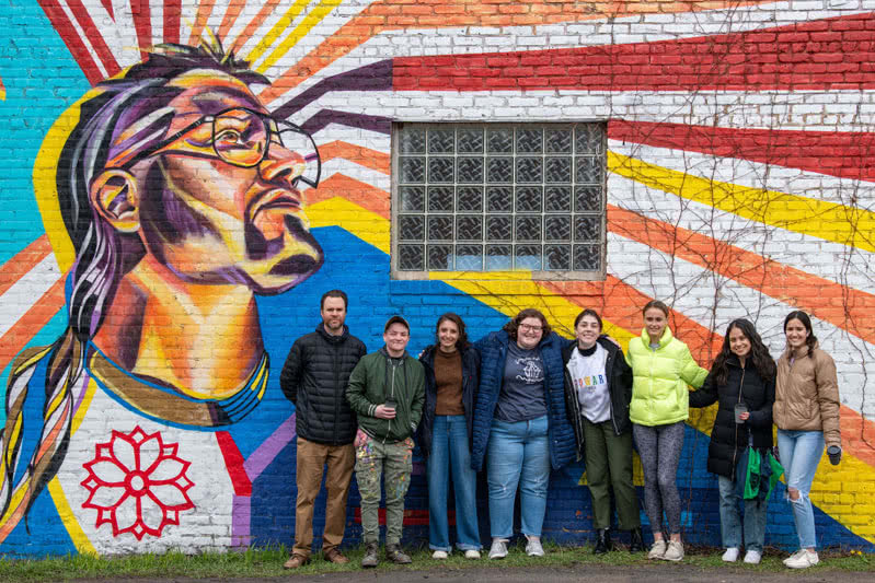 Colorful mural painted on the side of a building with 7 students and the painter standing in front.
