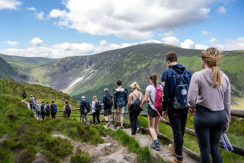 Students walking along a hilly path lined with a wooden post fence with rolling mountainous hills in the background.