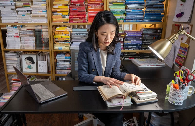 Woman sitting at desk reading a book with many multicolored and sorted by color books in a bookcase behind her.