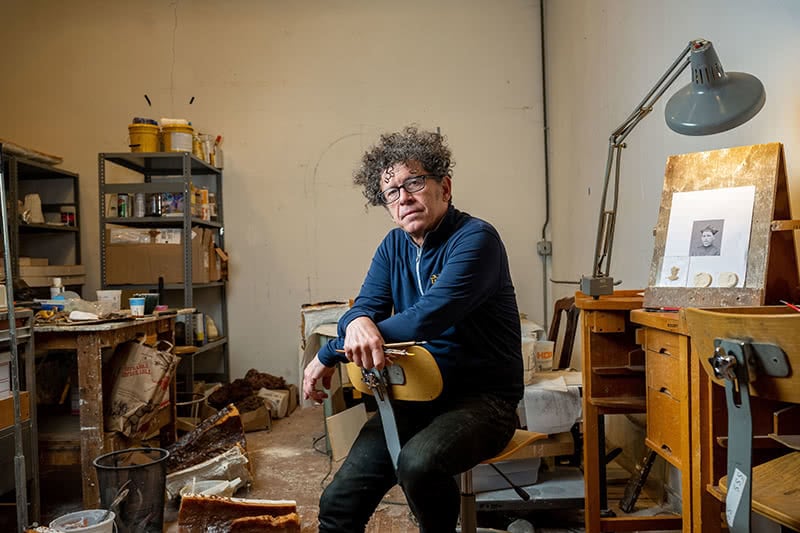 Miklos Simon sitting in his studio holding paint brushes, sitting in a chair backwards, looking at the camera.