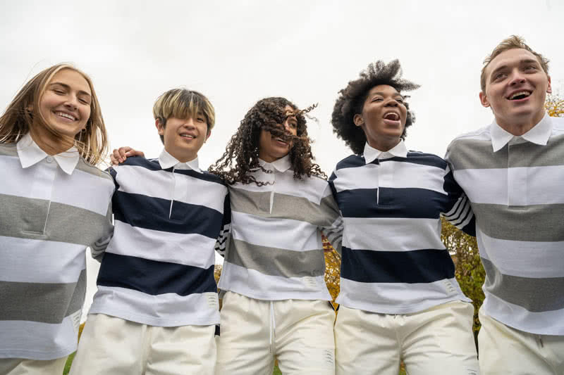 Students standing in arms looking down at the camera and laughing. Students are wearing matching striped shirts and solid white pants.