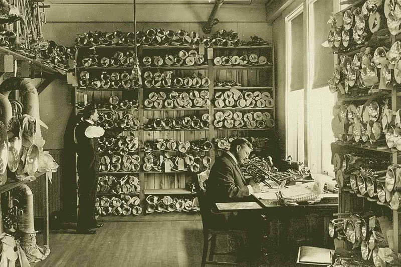 A yellowish archival photo of two white men in a room filled, floor to ceiling, with trumpets. One man sits at a desk holding a trumpet and takes notes. The other man stands against a wall of trumpets.