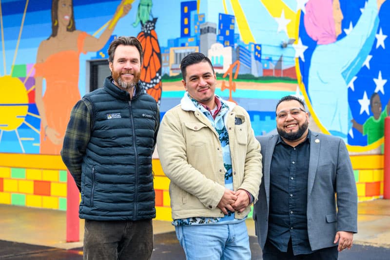 Three men stand in front of a mural created by Freddy Rodriguez at La Casa’s building on South Michigan St. in South Bend.