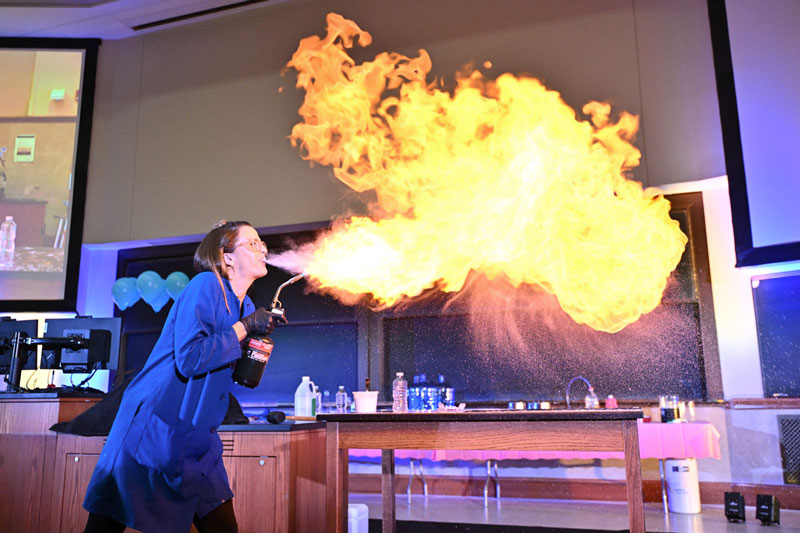 Kate the Chemist blows liquid out of her mouth over a flame, catching the liquid on fire. 