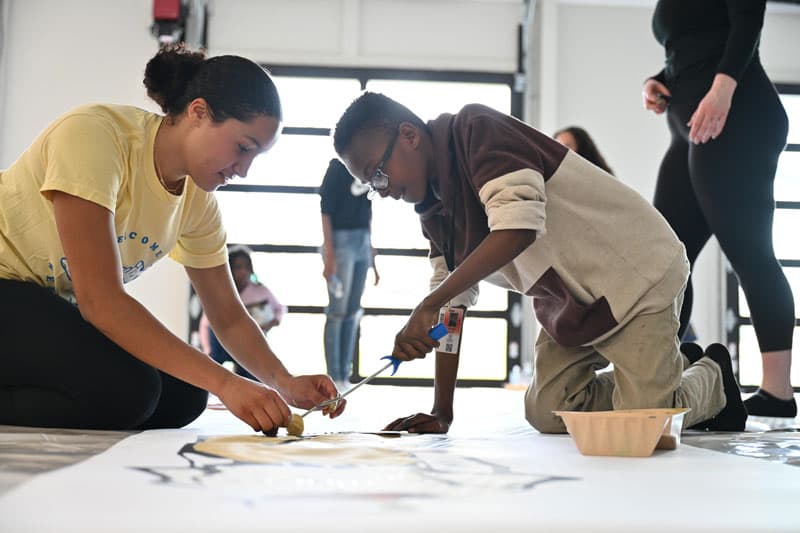 Two students sit on the floor and paint.