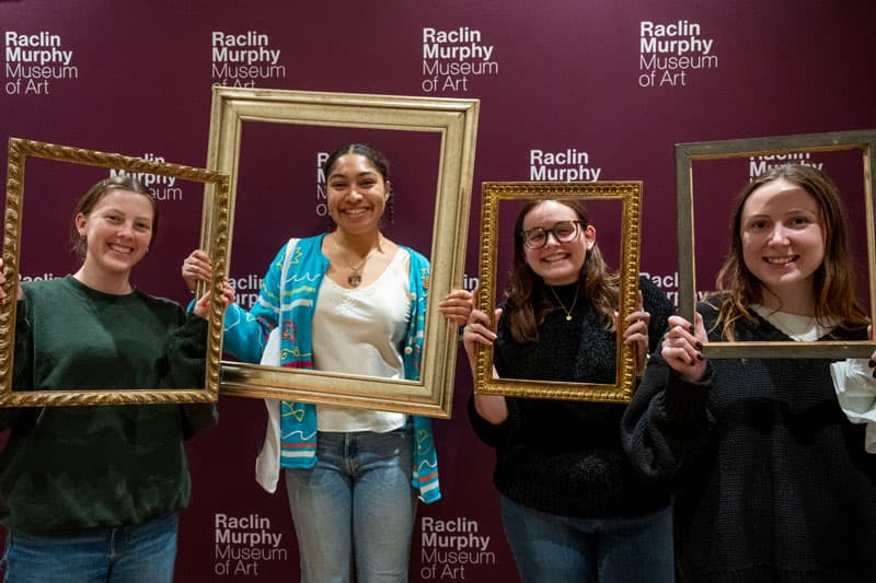 Four females stand in front of a purple backdrop while holding gol picture frames around their faces.