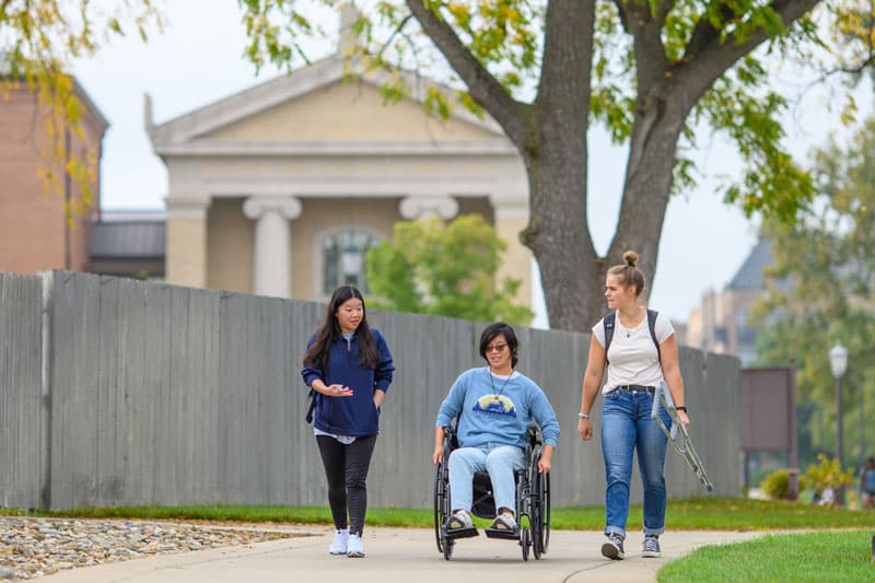 A student using a wheelchair rides down the sidewalk between two people, one who is carrying crutches.