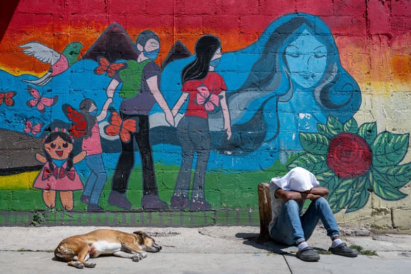 A migrant sits with his head between his knees next to a brightly painted wall.