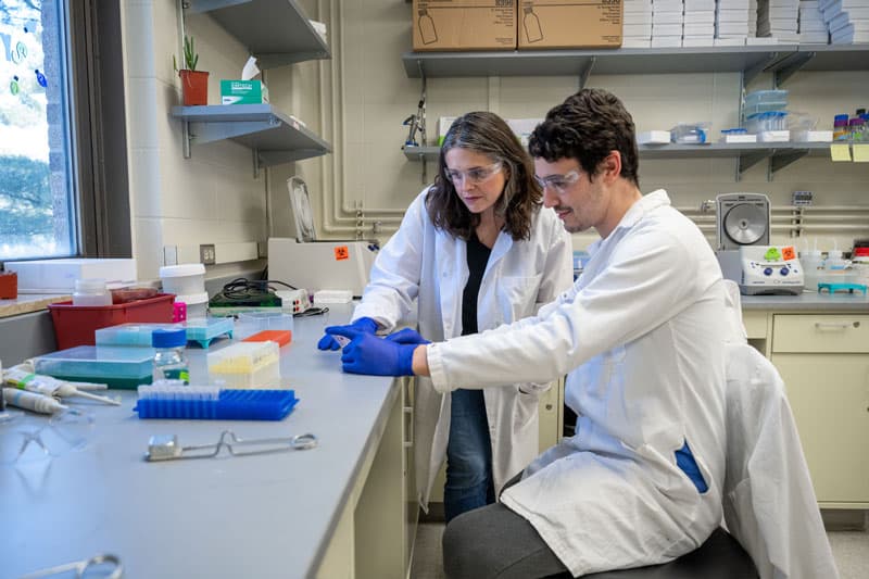 A male and a female in white lab coats look through a microscope.