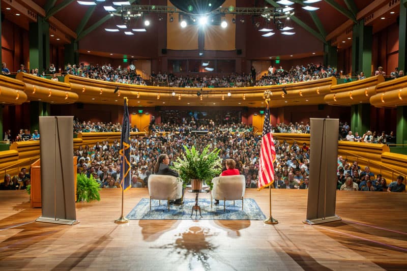 Two people sitting in chairs on a stage looking out into a packed auditorium. 