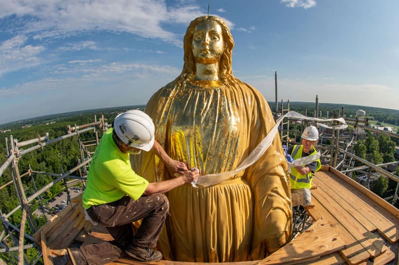 A man and a woman apply gold leaf to the statue of Mary on top of the Golden Dome.