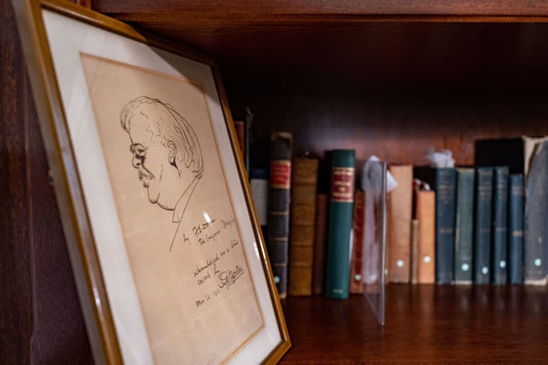 A caricature of GK Chesterton in a frame, sitting to next to a shelf with all his books.