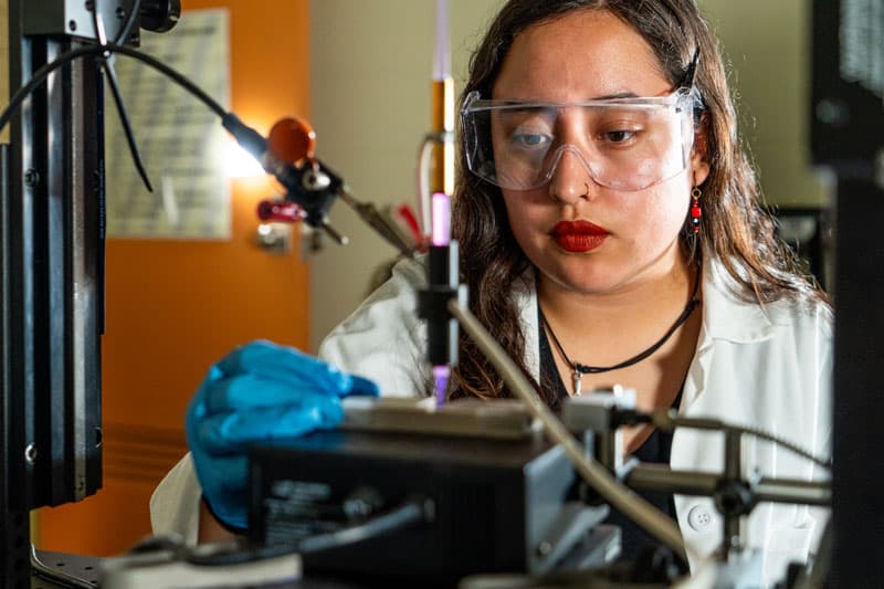 A female in a lab coat and safety goggles work at a machine.