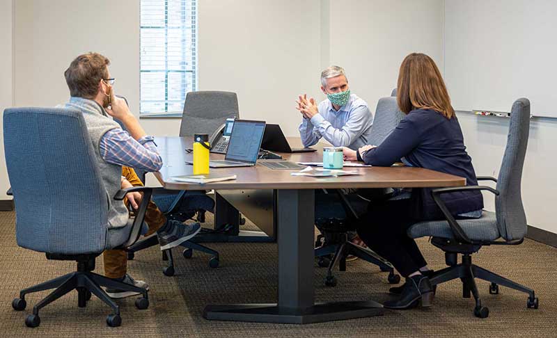 Three masked individuals sit at a conference table.