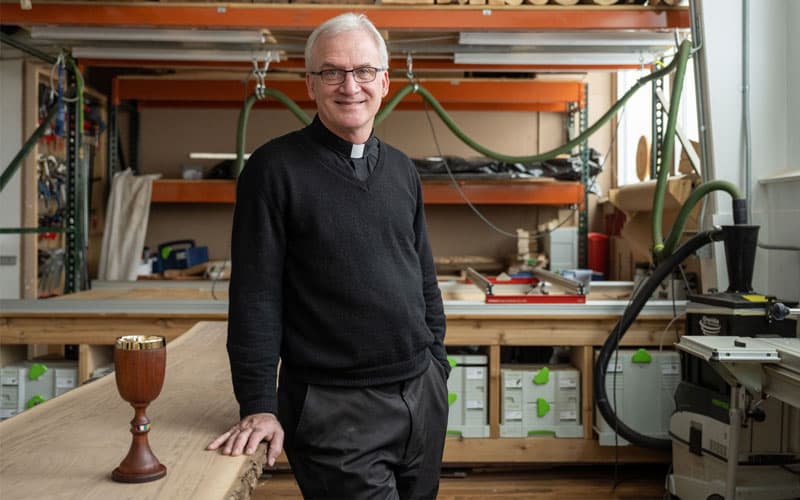 A photo of Fr. Dan Groody standing in his workshop with a wood chalice on the table next to him.