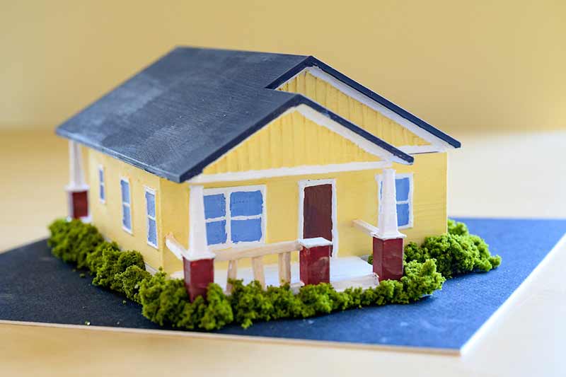A model of a model Habitat home on top of a graduation cap. The home is yellow, with a red door and dark gray roof. The home is surrounded by green bushes.