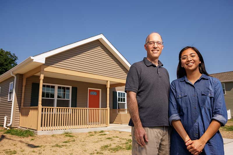 A male professor and female student pose for a photo in front of a one-story home with a bright orange door.