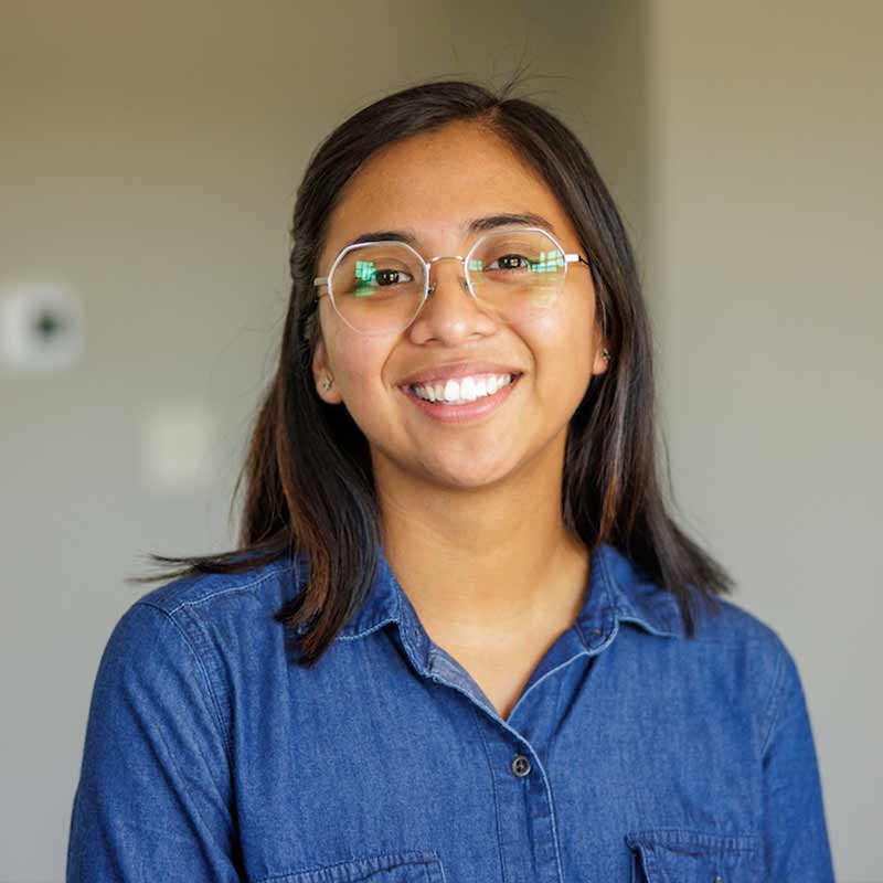 A headshot of Naomi Hernandez, a woman smiling, wearing glasses and a blue button up shirt.