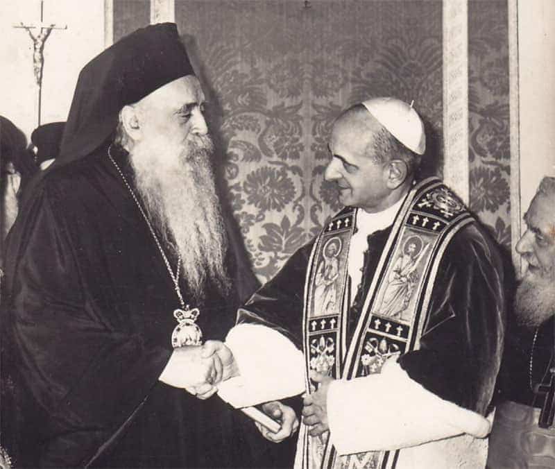 An old, washed out, black and white photo of a pope and an Orthodox shaking hands. One man has a long, white beard.