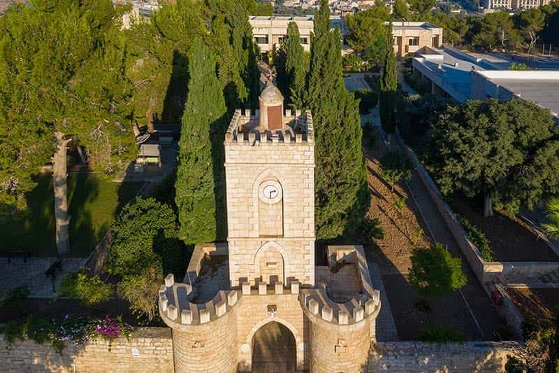 Aerial view of a stone tower with a clock and cross on top. Green cypress trees surround it, and a garden.