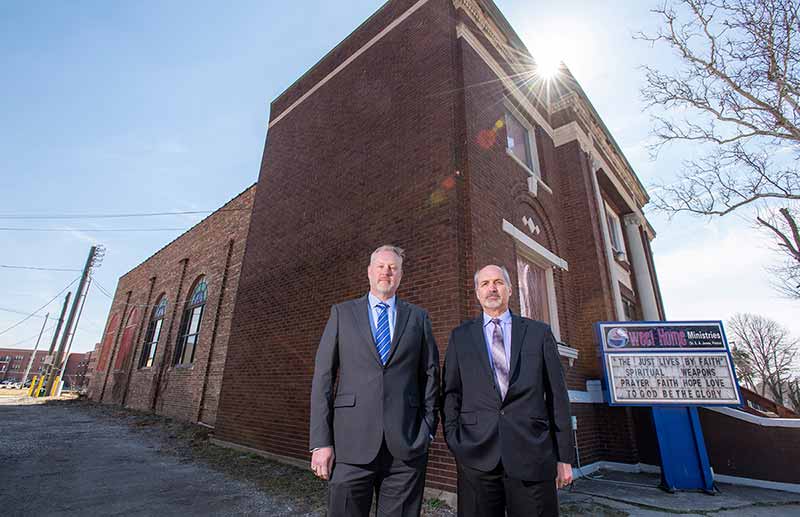 Two men stand in front of a brick church.