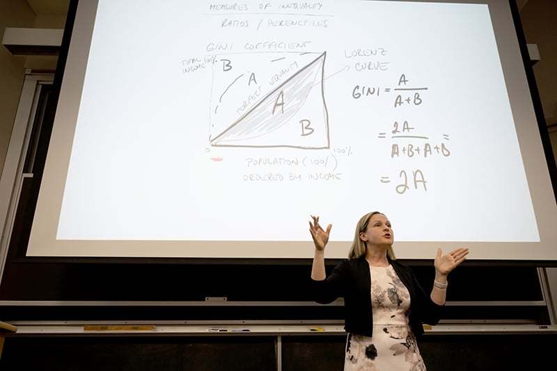 Dziadula standing at the front of a classroom, speaking with a projection of an equation behind her.