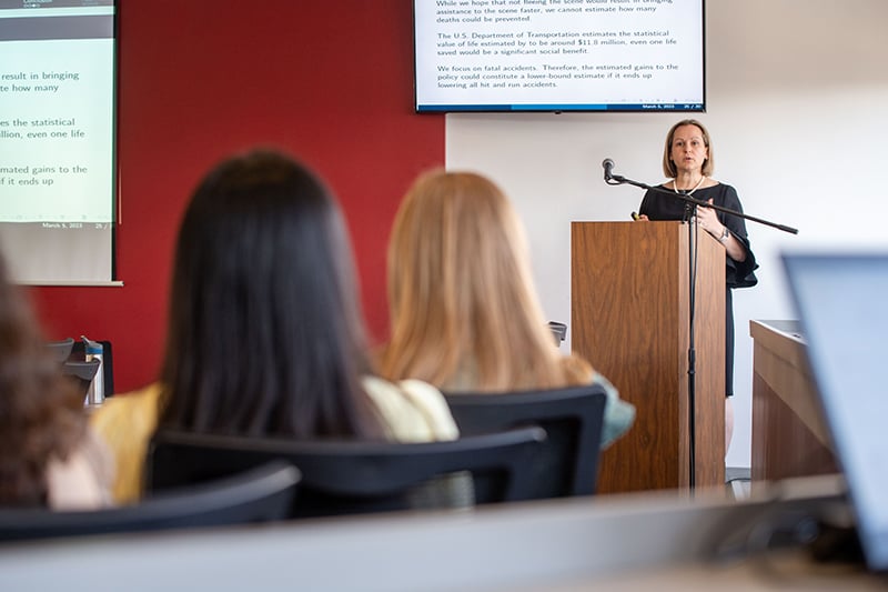 Dziadula standing at a podium with data projected behind her as she presents economic data at the APAEP conference.