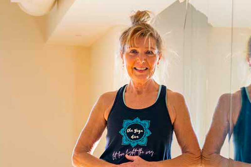 A woman wearing yoga gear in a yoga studio smiles to the camera.