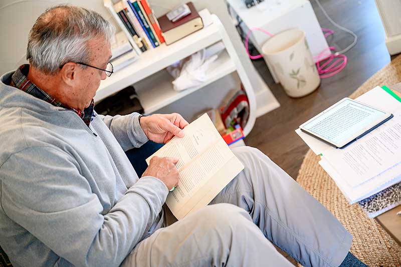 An older man reads a book on a couch in front of a coffee table with more books.