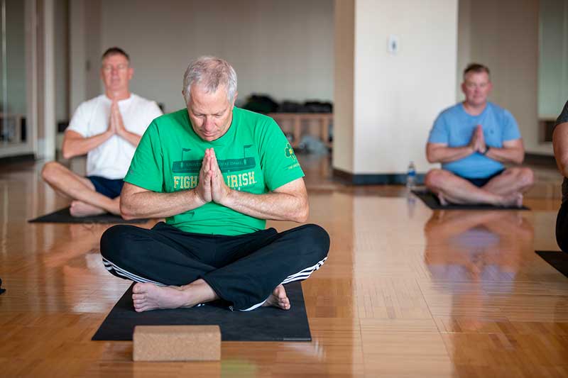 Men sit on yoga mats with their legs crossed and hands together at their chests.