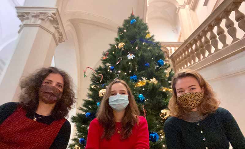 Three masked individuals sit in front of a Christmas tree.