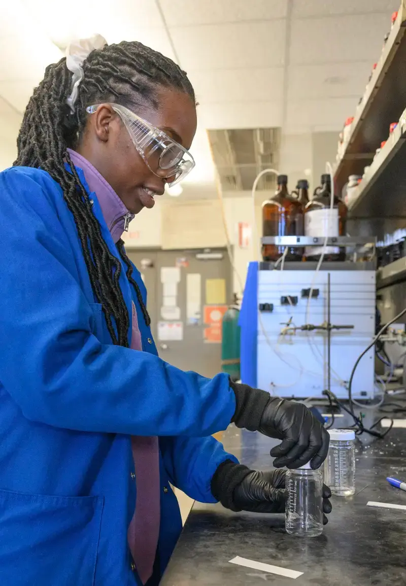 A black student wearing a blue lab coat stands at a table putting a lid on a glass beaker.