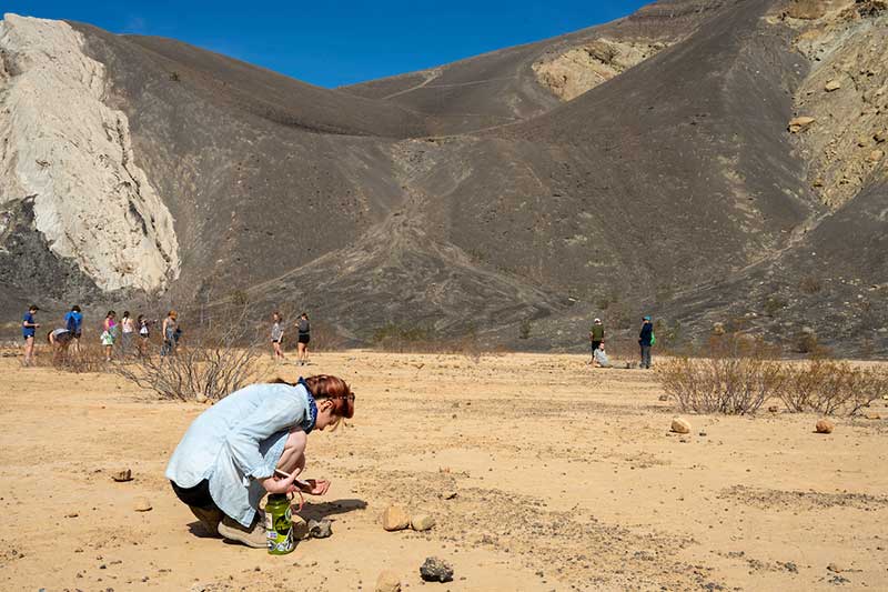 A female student crouches down and examines rocks in a crater. Behind her is a large moutain formation.