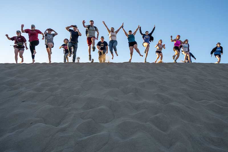 A group of students jump for a photo ontop of a sand dune.