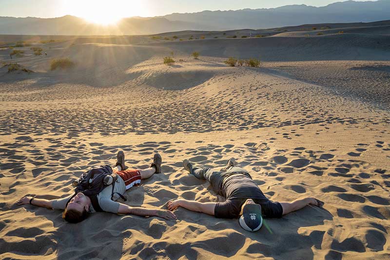 Two students lay on sand on their backs. The sun shines just above a sand dune in the distance.