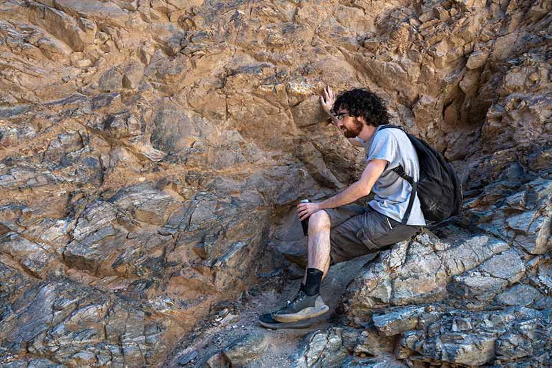 A male student sits on and sets his hand against a rock formation.