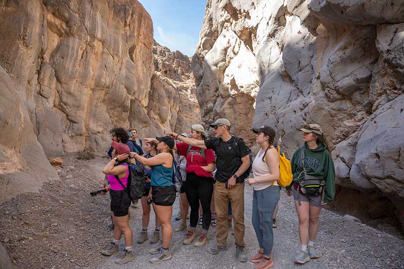 Inside narrow rock formations, a professor points to the side of a rock to a group of students.