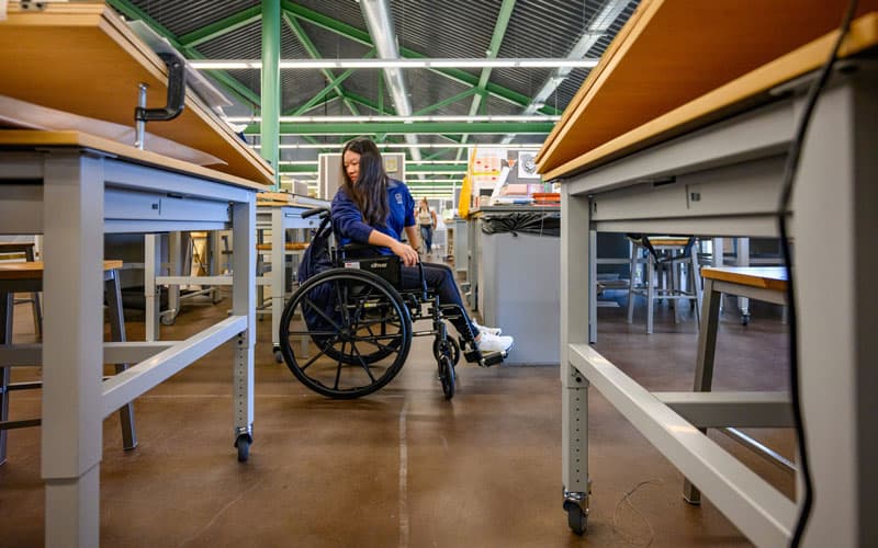A woman in a wheelchair is looking backwards, trying to figure out how to get through rows of desks.