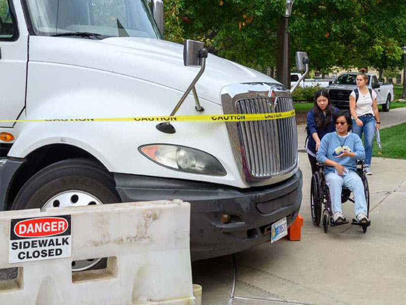 A student being pushed in a wheelchair is forced tp navigate an illegally parked truck on a sidewalk.
