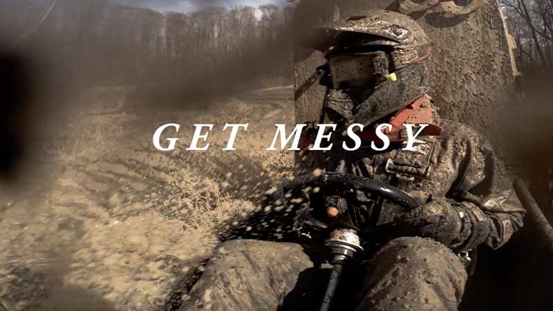 An individual drives a dune buggy through mud. Mud is splattered all over the diver and camera. 'Get Messy' text on top of the image.