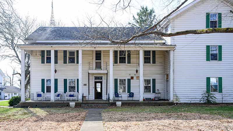 An older building with a large front porch where chairs and tables sit.