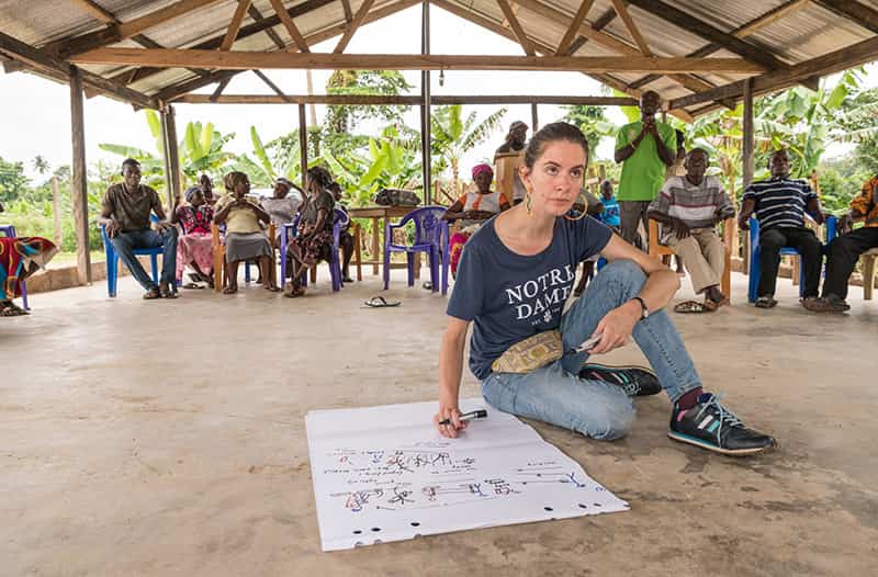 A woman, wearing a Notre Dame t-shirt, sits on the ground while drawing on a large piece of paper. Several people sit and stand behind her.