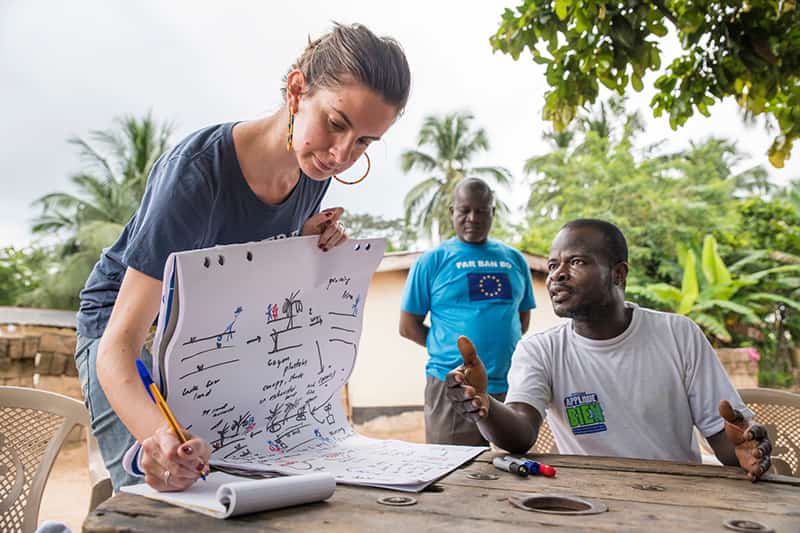 A man is expressive as he speaks to a woman while she writes notes on a pad of paper.