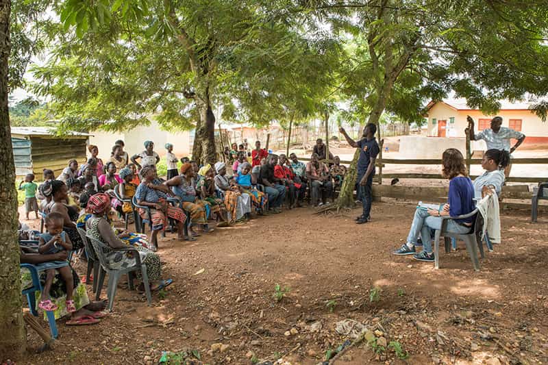 A man stands speaking to a large group of men and women and their children.