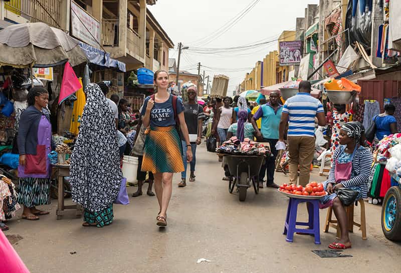 A woman walks down a busy market where people are selling fresh produce, colorful colthing, and many more items.