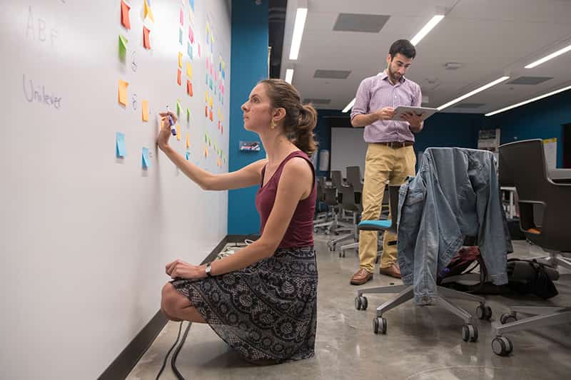 A woman crouched down leans on whiteboard and looks up at sticky notes. A man is in the background holding and looking at a tablet.