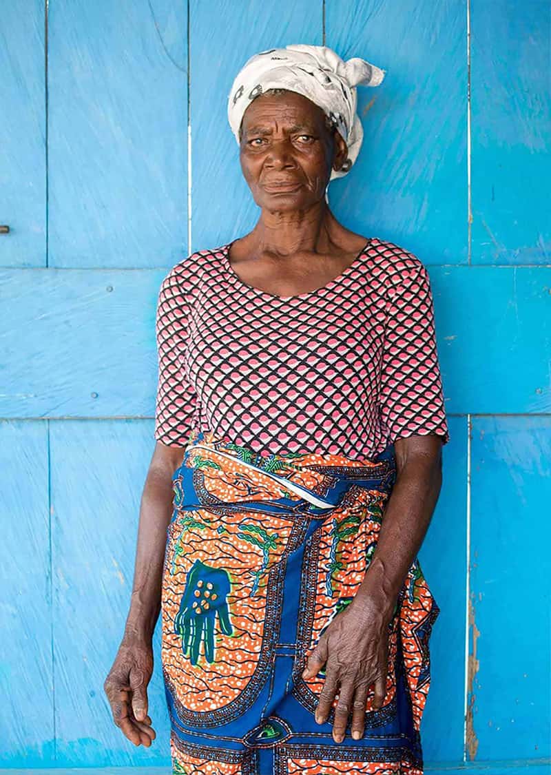 A Ghanaian woman wearing bright patterned clothes stands in front of an aqua colored wall.