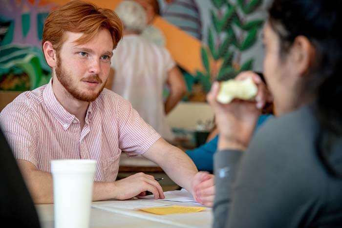 Brockman sits at a table with pen and paper, interviewing a woman.